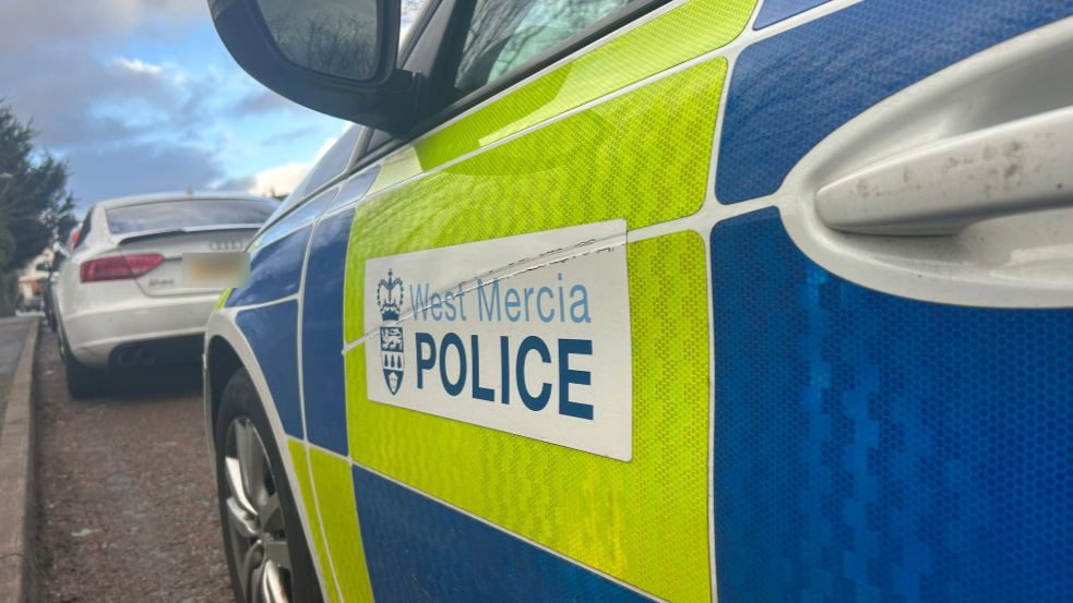 A close up image of a police vehicle's door. The door is predominately white with yellow and blue high-vis stickers on the side as well as a white sticker that reads "West Mercia Police".
