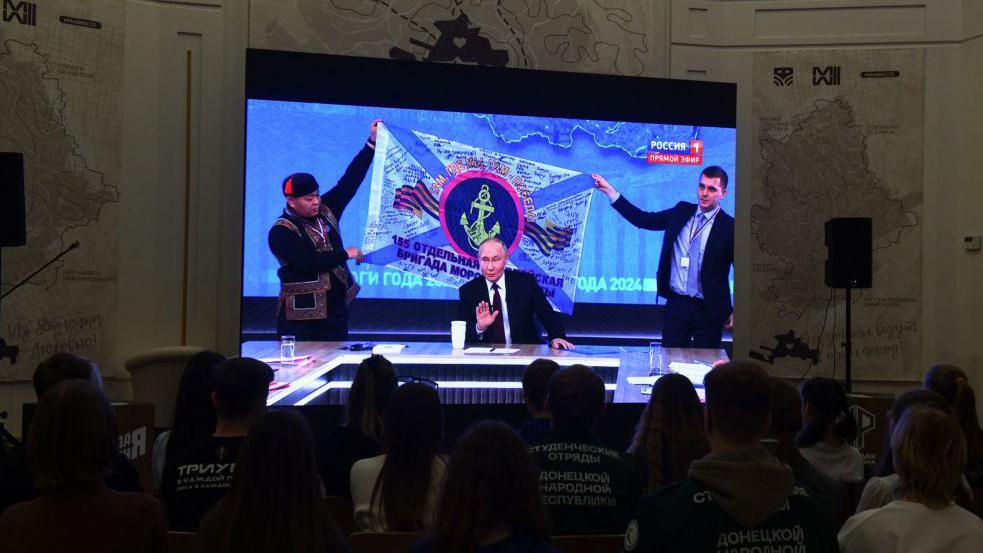 Putin on a TV screen, behind him staff members hold-up a flag he said was given to him by Russian marines fighting in Kursk 