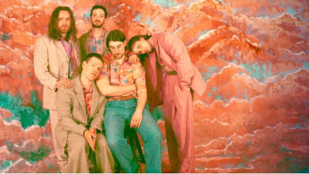 The five members of Cousin Kula band leaning on each other affectionately in front of a pink and blue painted cloud background. They are all wearing colourful baggy suits with retro style shirts.