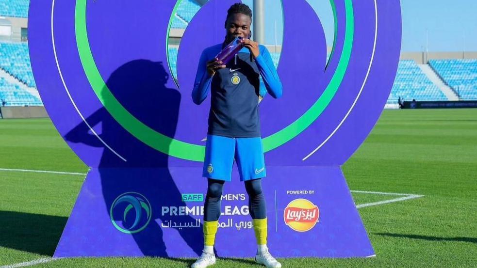 Clara Luvanga stands in training gear on a pitch in front of a promotional hoarding and holds up an award