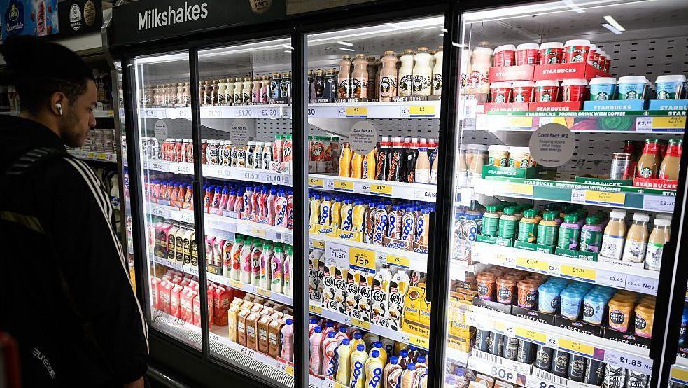 A man with dark hair stands in front of a large supermarket fridge containing shelves of milk-based drinks, such as flavoured milkshakes and milky coffees like lattes, cappucinos and iced coffees.