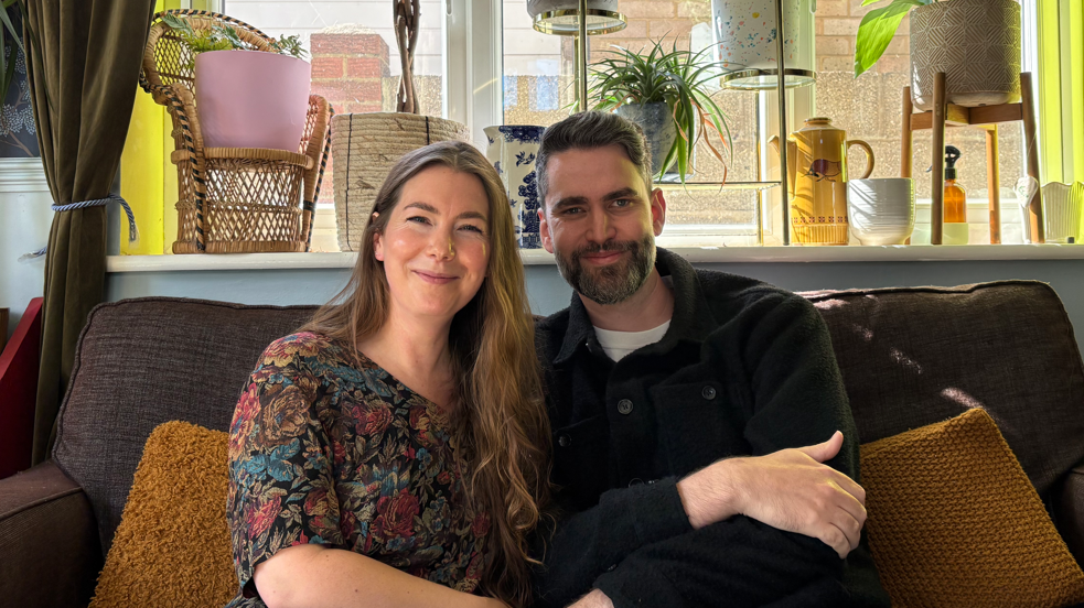 Jenny and Joe are sitting closely on a couch in a cozy, well-lit living room. The background features homey details like plants, wicker baskets, and decorative items on a windowsill. Their arms are linked as they both smile at the camera. Jenny has long wavy light brown hair and is wearing a black top with red, green, and blue floral detailing. Joe has dark hair and some grey in part of his facial hair. He is wearing a white top and a black jacket.
