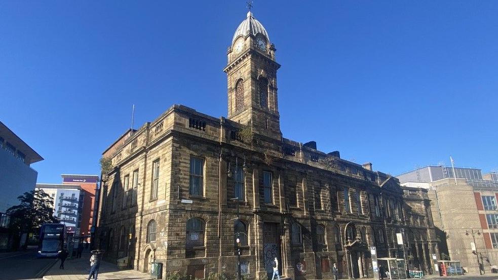 An exterior image of the former Sheffield Town Hall with a blue sky. A two-storey neoclassical-style building with a clock tower rising above it.