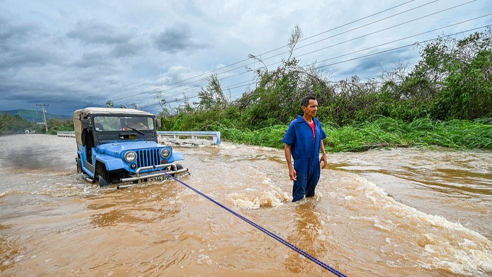 A man wearing a blue jumpsuit stands in brown floodwater beside a bright blue car that is partially submerged. The vehicle is being pulled or secured with a rope attached to the front bumper.