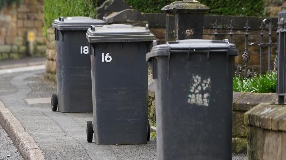 A stock image of wheelie bins on a pavement outside of a house. There are three black colour wheelie bins. The two towards the back of the image have number 16 and 18 on them.