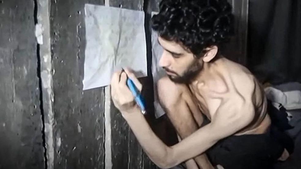 Evyatar David holds a pen and writes on a paper as he crouches, emaciated, in a tunnel in Gaza