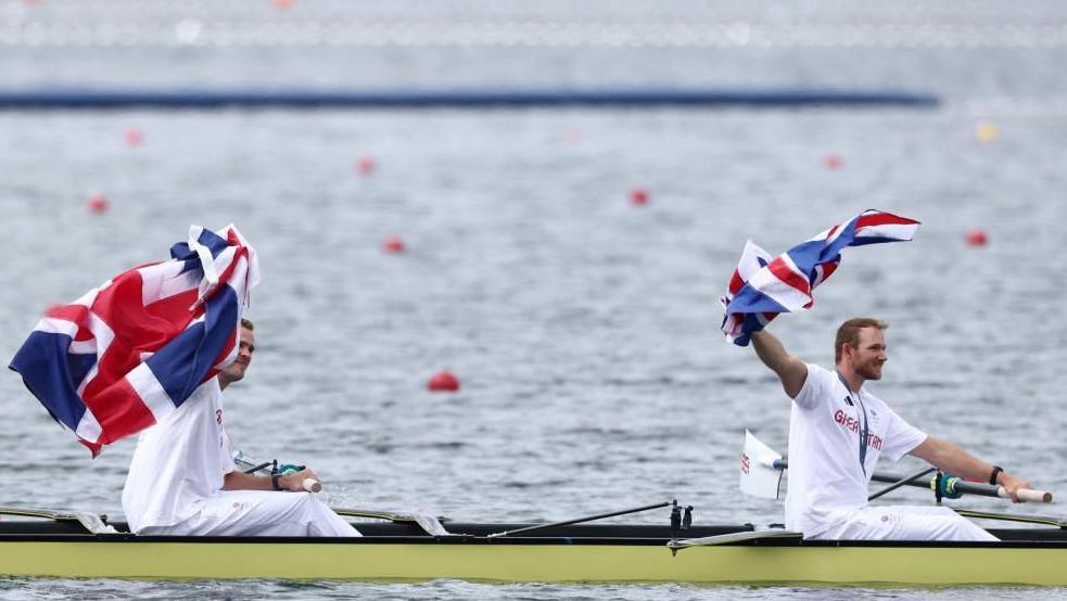 James Rudkin and Charles Elwes holding flags in a boat