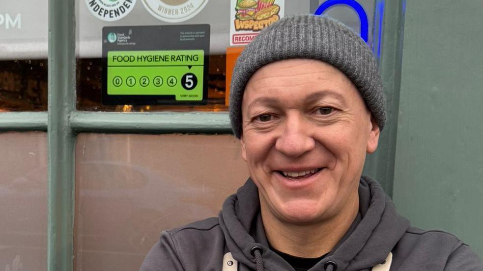 Marc Ward stands on the right of the image, looking towards the camera and smiling. He is wearing a grey hat and hoodie. He is standing in front of a building next to a window displaying a five-star food hygiene rating sign.