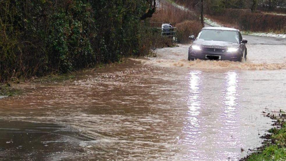 A grey Land Rover with snow on its bonnet and roof drives through floodwater which is about two feet high
