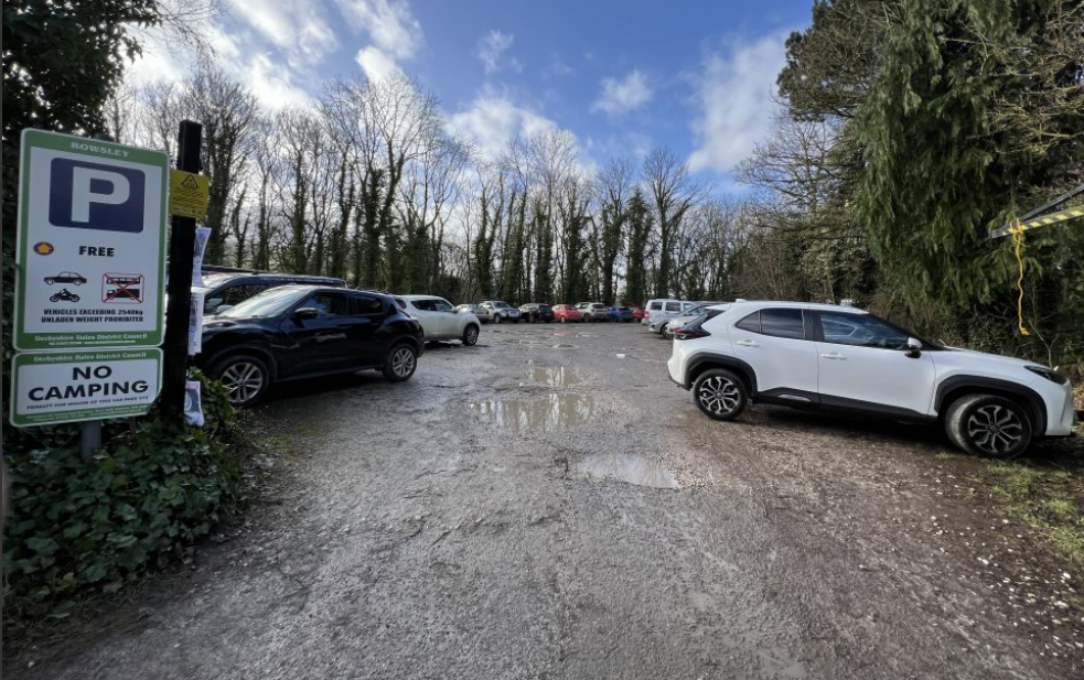 car park with puddles on the ground with several cars parked, surrounded by trees 