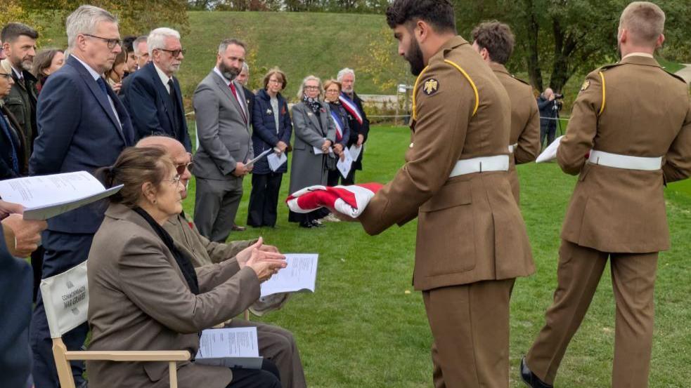 A woman, the descendant of one of the four Grimsby soldiers, is handed a Union flag, folded into a rectangles, in the Commonwealth War Graves Commission cemetery. The woman is sitting on a chair, with an order of service on her lap, as she is presented with the flag by a serving soldier.