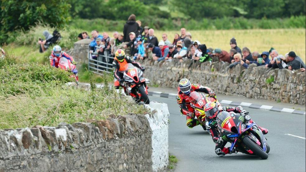 Racers in colourful leathers leaning in for a bend. There are stone walls either side of the road and spectators watching on in the background.