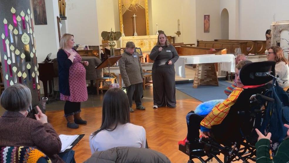 Two women and a child stand at the front of the room performing with the rest of the group watching along.  They are stood on a wooden floor with a piano behind the two women