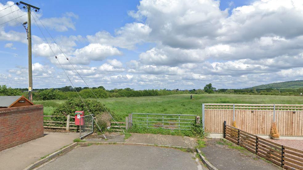 A field near Sinderberry Drive in Northway. There is a gate blocking the entrance and trees and hills can be seen in the distance