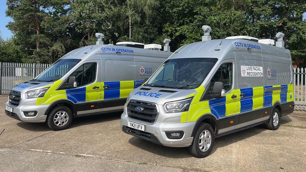 Two grey live facial recognition vans covered in police signage. They are next to each other and static. They are parked on a concrete with a metal fence behind them. 