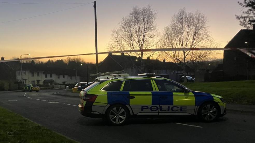 A yellow blue and white police car sits in the middle of the road at a security alert in Twinbrook