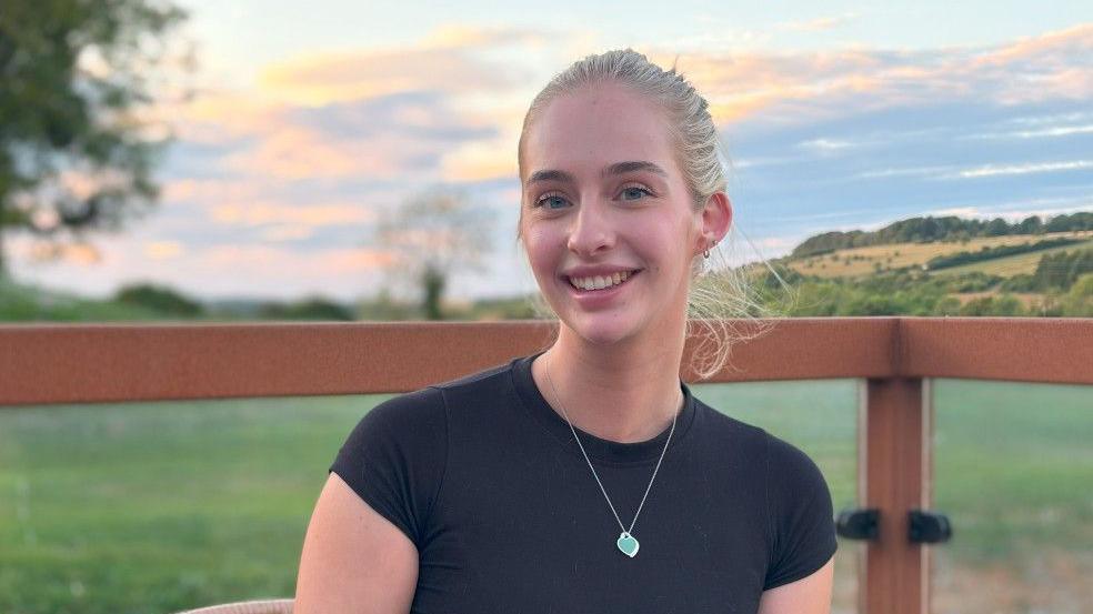 Young woman with blonde hair, wearing a black short-sleeved shirt and green pendant necklace. She is sitting in countryside with fields and trees in the background.