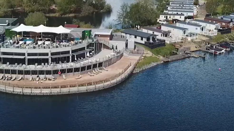 An aerial image of Tattershall Lakes Country Park shows a lakefront with several long, thin static caravans on the shore, and a couple of small boats moored up. There's also a large communal area and beach with sunloungers set out and a large white parasol offering some shade.