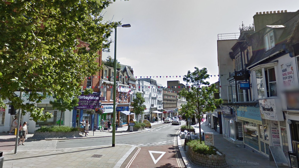 A road junction with a number of shops either side of a road with trees either side of the road.