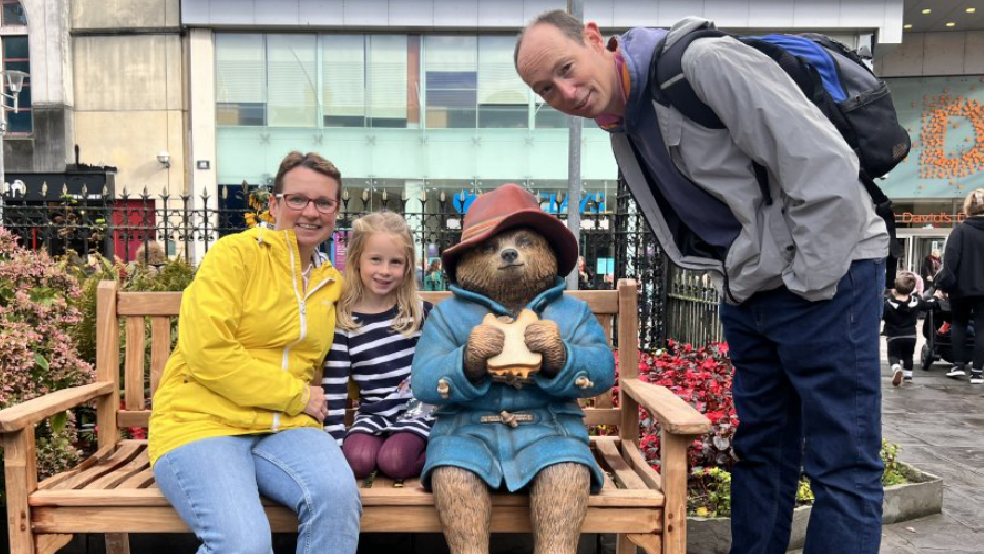 Parents Karen and Graham with their daughter Orla, sitting next to the Paddington bear statue. 