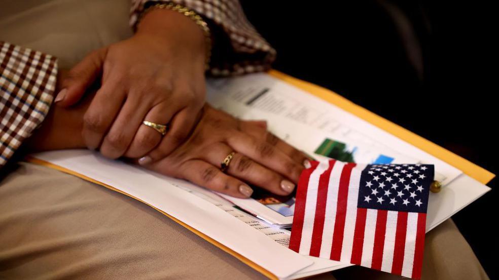 A black woman's hands, with pink manicured nails and a two rings, are crossed i her lap on top of a small stack of papers and a small US flag