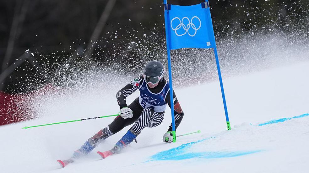 Lasse Gaxiola skies past a blue Olympic sign, sending flurries of snow into the air.