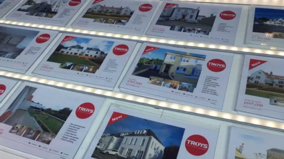 An estate agent window with pictures of different properties for sale. In a red circle in the top right corner of each of the adverts, it has the words 'TROYS'.