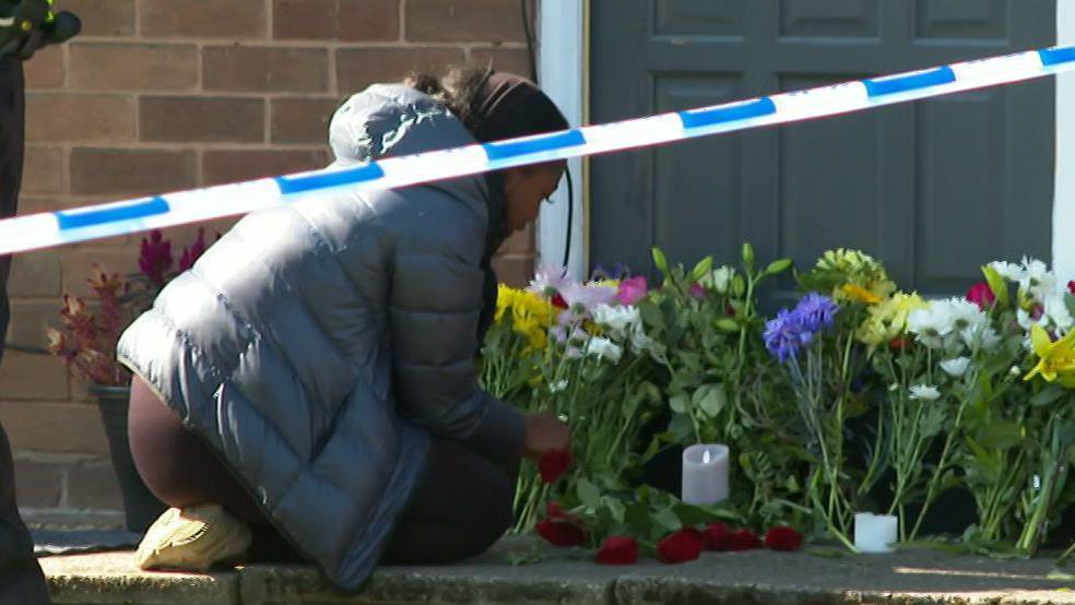 A woman kneels and arranges red roses around a candle as floral tributes are left for Jahziah Coke.