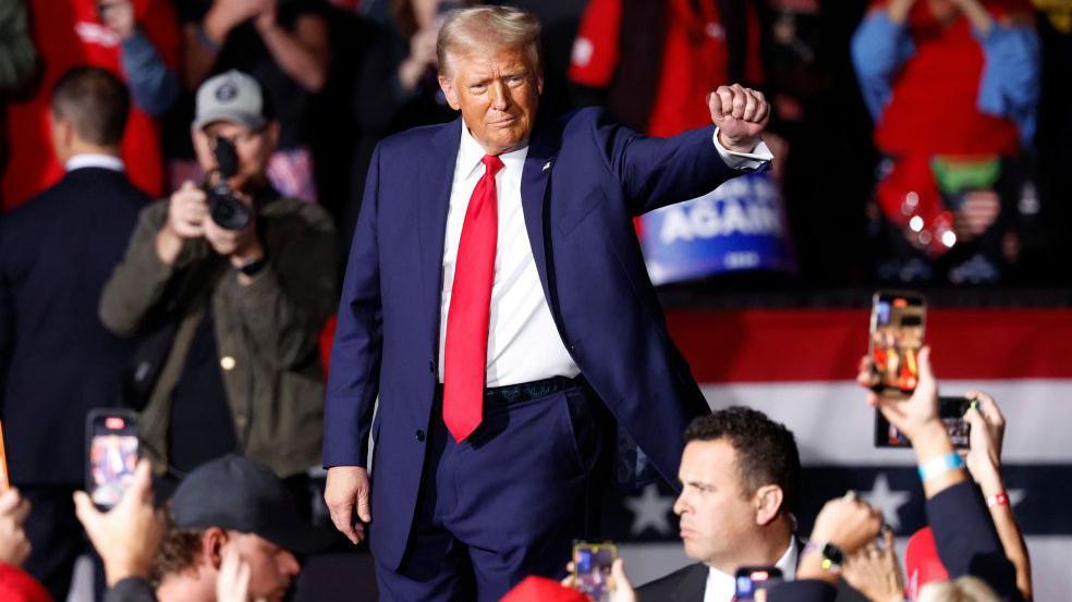 Donald Trump walks through an arena crowd with his right fist raised out. Several mobile phones are photographing and videoing him.  