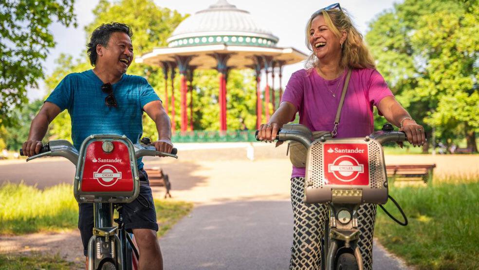 A man, in a blue top and shorts, and a woman, in black and white trousers and pink top, are cycling on red Santander rental bikes along a paved path in a sunny park. Behind them is a pavilion, surrounded by green trees and grass.