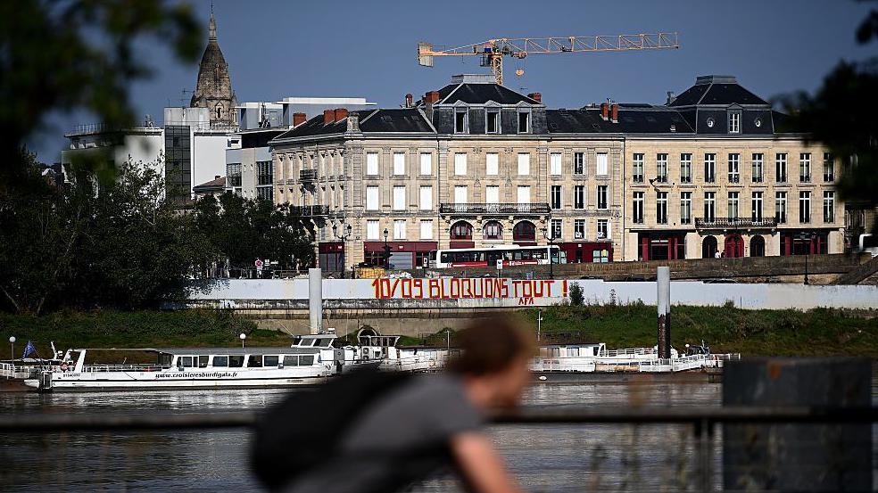 A cyclist pedals past graffiti reading 'September 10 Let's block everything' on the wall of a construction site on a river in Bordeaux