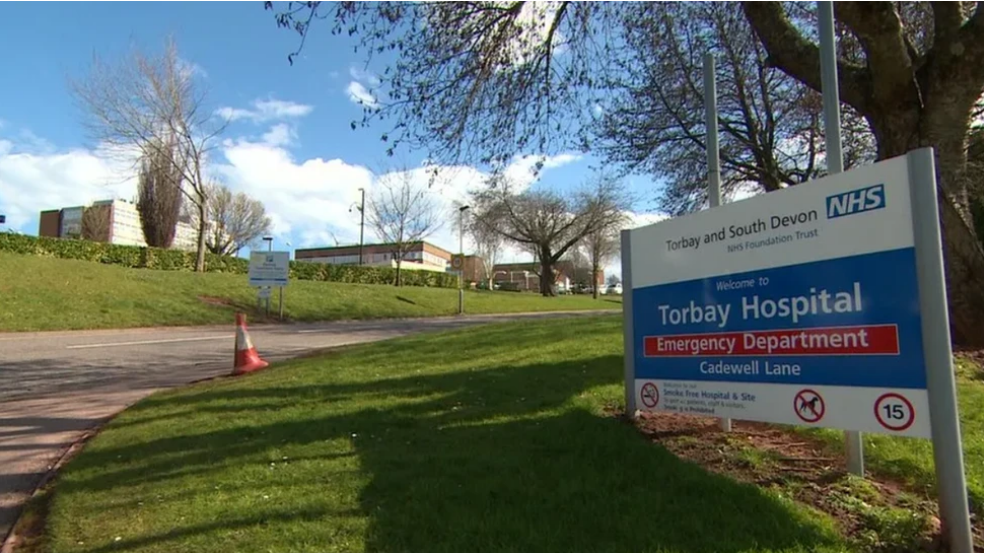 On the left is a white and blue sign with 'Torbay and South Devon NHS Foundation Trust' at the top and below 'welcome to Torbay Hospital Emergency Department Cadewell Lane'. In the background there is grass and a road leading the hospital with trees.