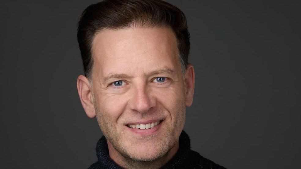 Headshot of Andrew Husband smiling at the camera. He has brown hair and is wearing a black polo jumper with a black background.