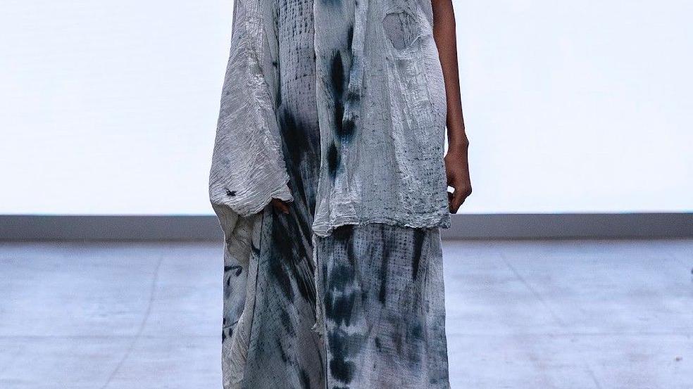 A close up of the lower portion of a design. A model is walking down the catwalk wearing a pale blue tattered sheet with darker blue stains and splashes.