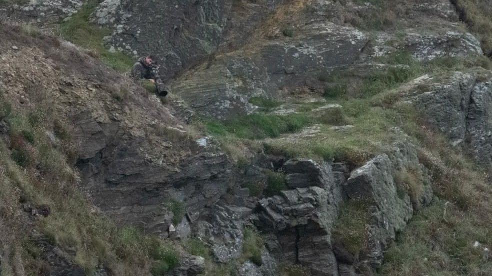 A man using a telescopic camera lens on a clifftop to take a photograph of something below. He is wearing a camouflage jacket to blend in to the green and grey of the rock and grass behind him.