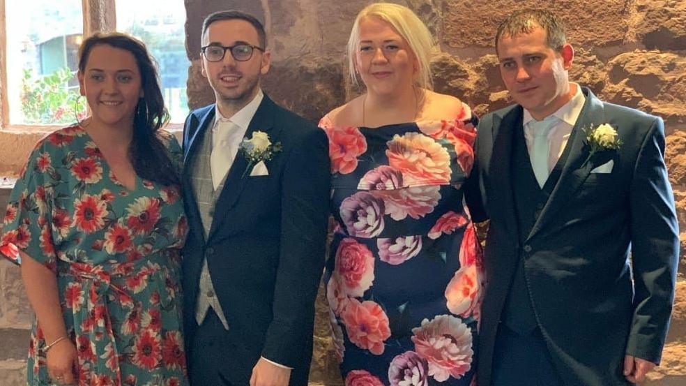 The four siblings are standing next to each other in formal attire at what appears to be a wedding celebration. Catherine and Emma have floral dresses while Luke and James are wearing morning suits with a cream rose buttonhole.