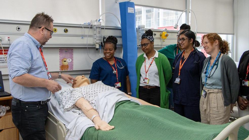 A man and a group of women stand over a mannequin lying in a hospital bed. There are five women standing over the mannequin.