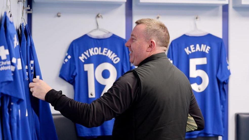 Tony Sage is standing in the dressing room at Goodison Park. He is in front of Everton players' blue shirts, hanging up on coat hangers.