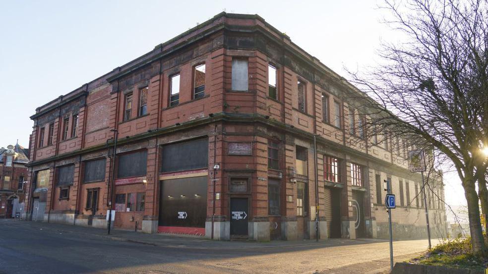 An old Victorian red brick building viewed from the corner, with empty roads feeding off to left and right. The building appears derelict with some windows and doors boarded up.