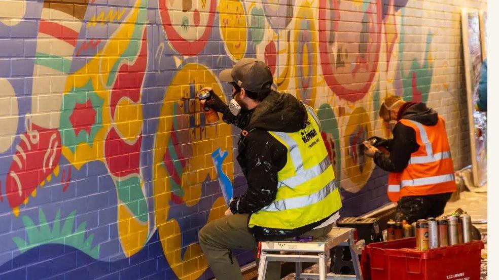 Two people in high viz tabards are sitting down and painting a wall with vibrant colours and designs. 