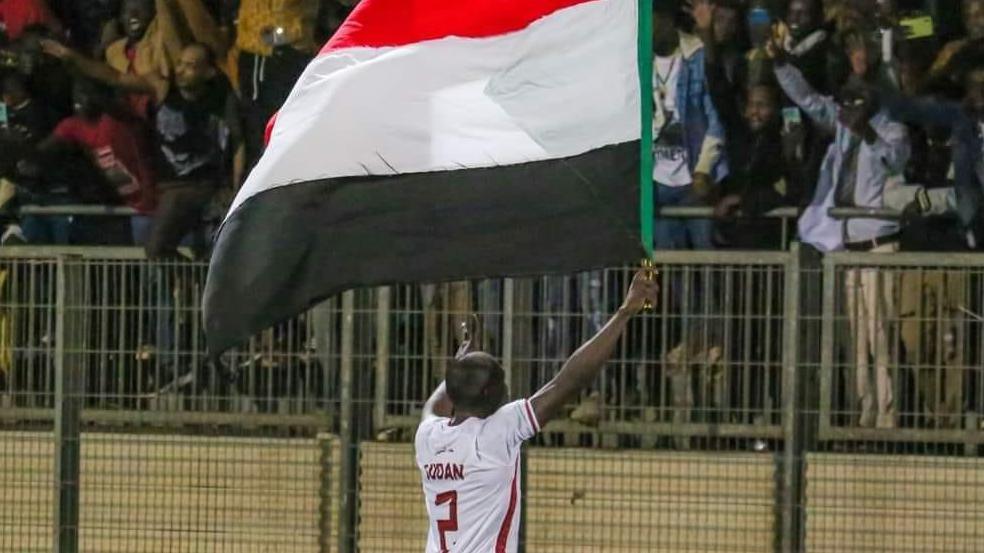 A footballer wearing a white Sudan shirt with his back to the camera raises a red, white and black Sudan flag in front of a crowd of cheering supporters as they celebrate a victory