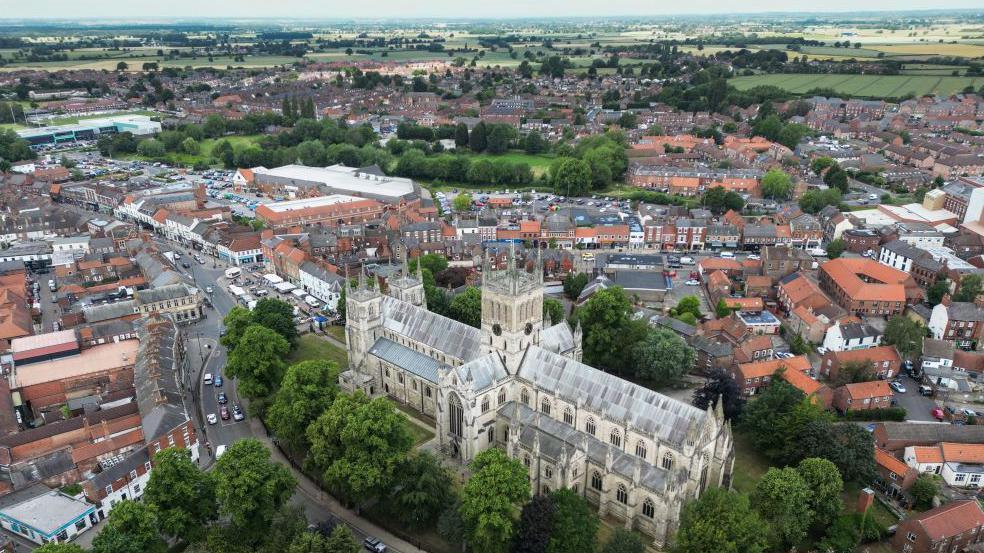 A large cathedral sits in the foreground surrounded by trees in the this view from above. Further afield is a town with many red tiled roofs.