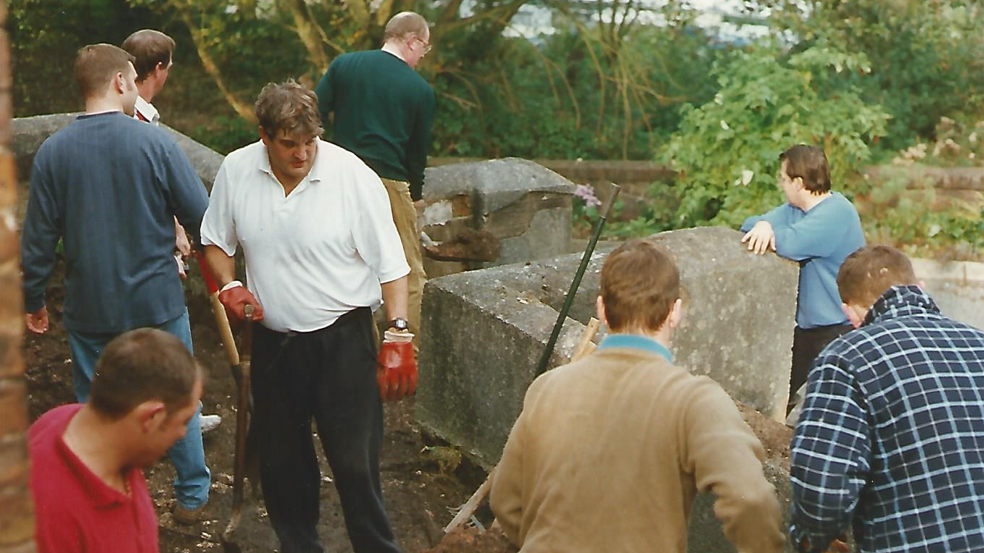 Eight men can be seen working on the groundworks of the chapel. They are clearing ground. Many have their back to the camera. Large stonework can be seen by where the men are working.