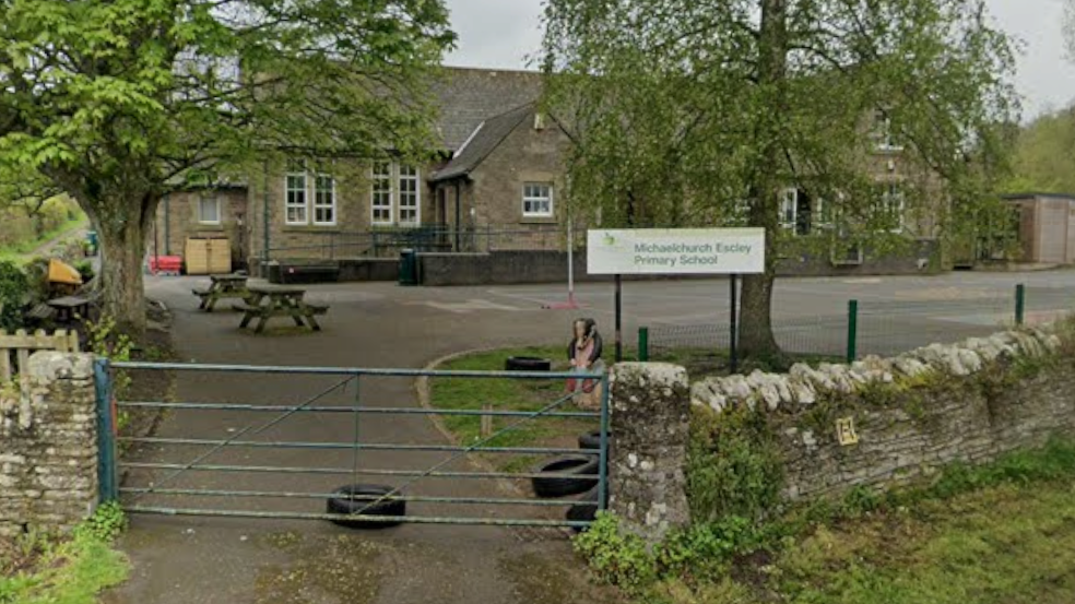 A large stone building stands behind a stone wall and a green metal gate. There are benches in the grounds, and trees, with a sign at the entrance saying Michaelchurch Escley Primary School.