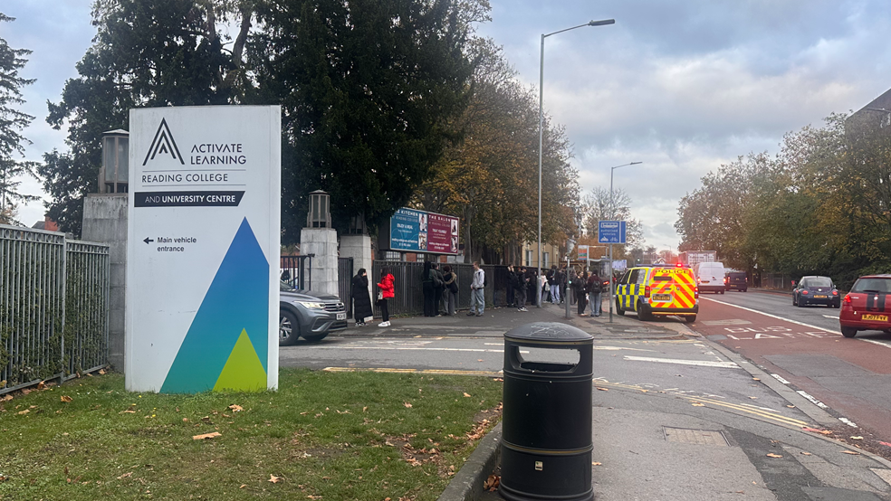A police van parked outside Reading College, with students milling around and a sign on the left for the college.