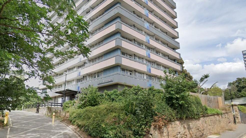 Nauls Mill House in Coventry, a tower block with a wall and garden area in front of it, containing shrubs, and a road approaching it with diamond-shaped markings and a yellow bollard.