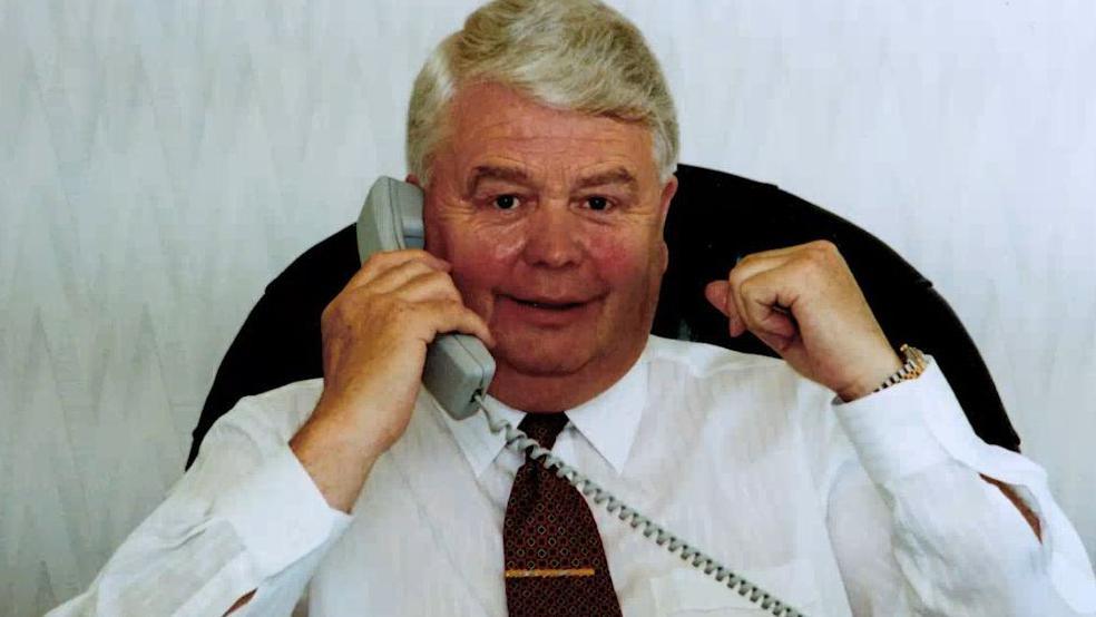 Bernard O’Malley with grey hair smiling as he holds a telephone to his ear sitting on an office chair wearing a white shirt with a dark red tie and gold tie pin and silver and gold watch