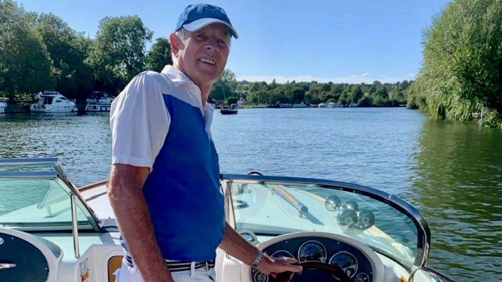 An elderly man in a blue cap and a blue and white polo shirt, standing in the cockpit of a boat with a hand on the steering wheel, smiling at the camera with a tree-lined river in the background