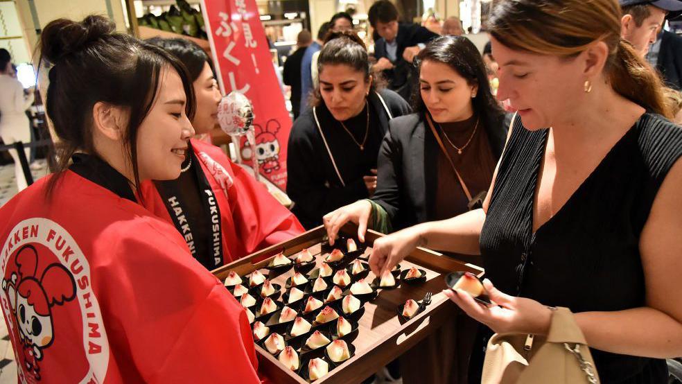 A group of women pick peaches from a tray being held by two women in Japanese kimonos