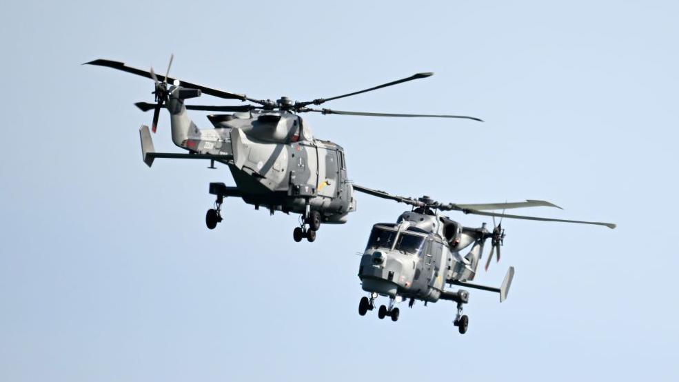 Royal Navy Black Cats display team in two Lynx helicopters, which are painted with a camouflage design, the appear to be flying towards one another which is part of the team's aerial display.